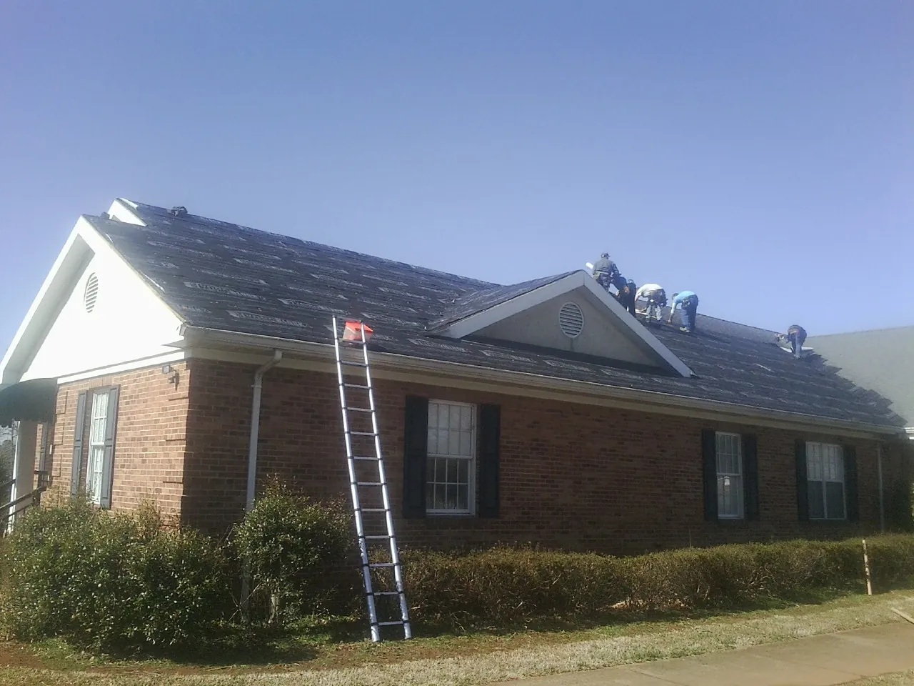 Crew tearing shingles off a brick ranch roof with a ladder set — Emergency Roof Repair work by our crew in Plainview