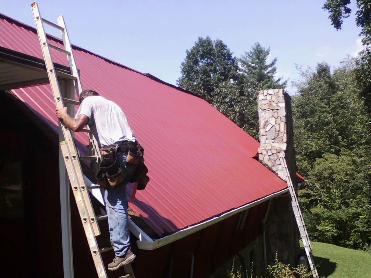 Roofer on a ladder beside a red metal roof and stone chimney — Soffit & Fascia Repair by Roofing Brothers in Plainview